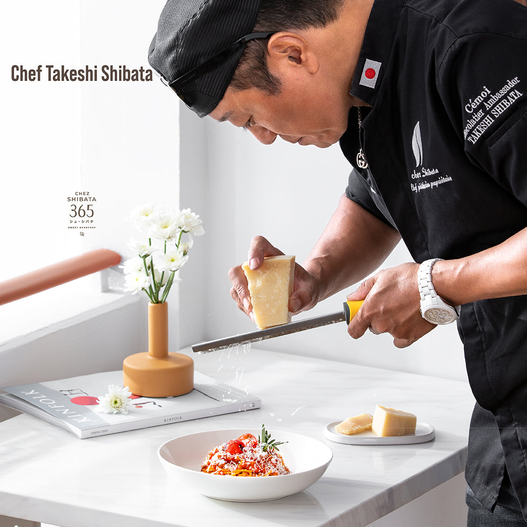 Chef Takeshi Shibata grating Parmesan cheese over a freshly prepared pasta dish at Chez Shibata 365. The setting is clean and elegant, highlighting the chef’s precision and craftsmanship.