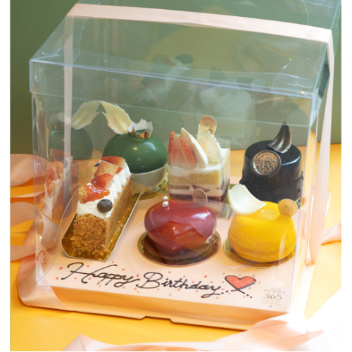กล่องของขวัญเค้กแบบเลือกเอง (Craft Your Own Gift) บรรจุเค้ก 5 ชิ้นในกล่องอะคริลิกใส ผูกริบบิ้นสีชมพู ด้านในมีเค้กหลากหลายชนิด เช่น Mille Feuille, Strawberry Shortcake, Yuzu Tart, และเค้กมูสตกแต่งอย่างประณีต ฐานกล่องมีข้อความ “Happy Birthday” พร้อมหัวใจ พื้นหลังเป็นโทนสีเขียวและเหลือง ดูหรูหราและอบอุ่น