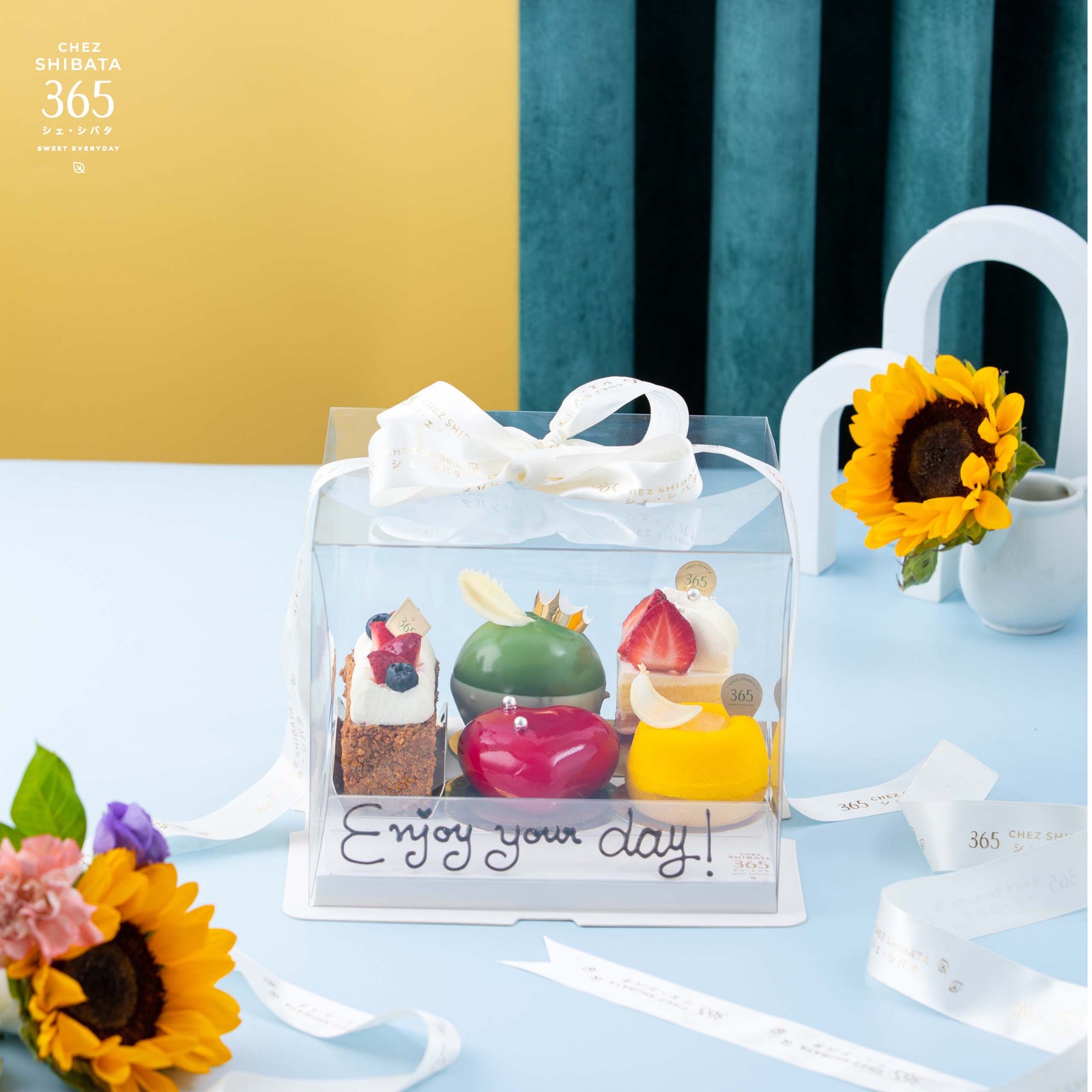 กล่องของขวัญเค้ก 5 ชิ้นบรรจุในกล่องใส พร้อมริบบิ้นสีขาว ด้านในประกอบด้วยเค้กหลากหลายสไตล์ ได้แก่ Mille Feuille, Matcha Queen, Strawberry Shortcake, Yuzu Tart และ Coeur des Bois ตกแต่งอย่างประณีต วางบนโต๊ะสีฟ้า ประดับด้วยดอกทานตะวัน เพิ่มความสดใสและอบอุ่น ข้อความบนกล่องเขียนว่า “Enjoy your day!”