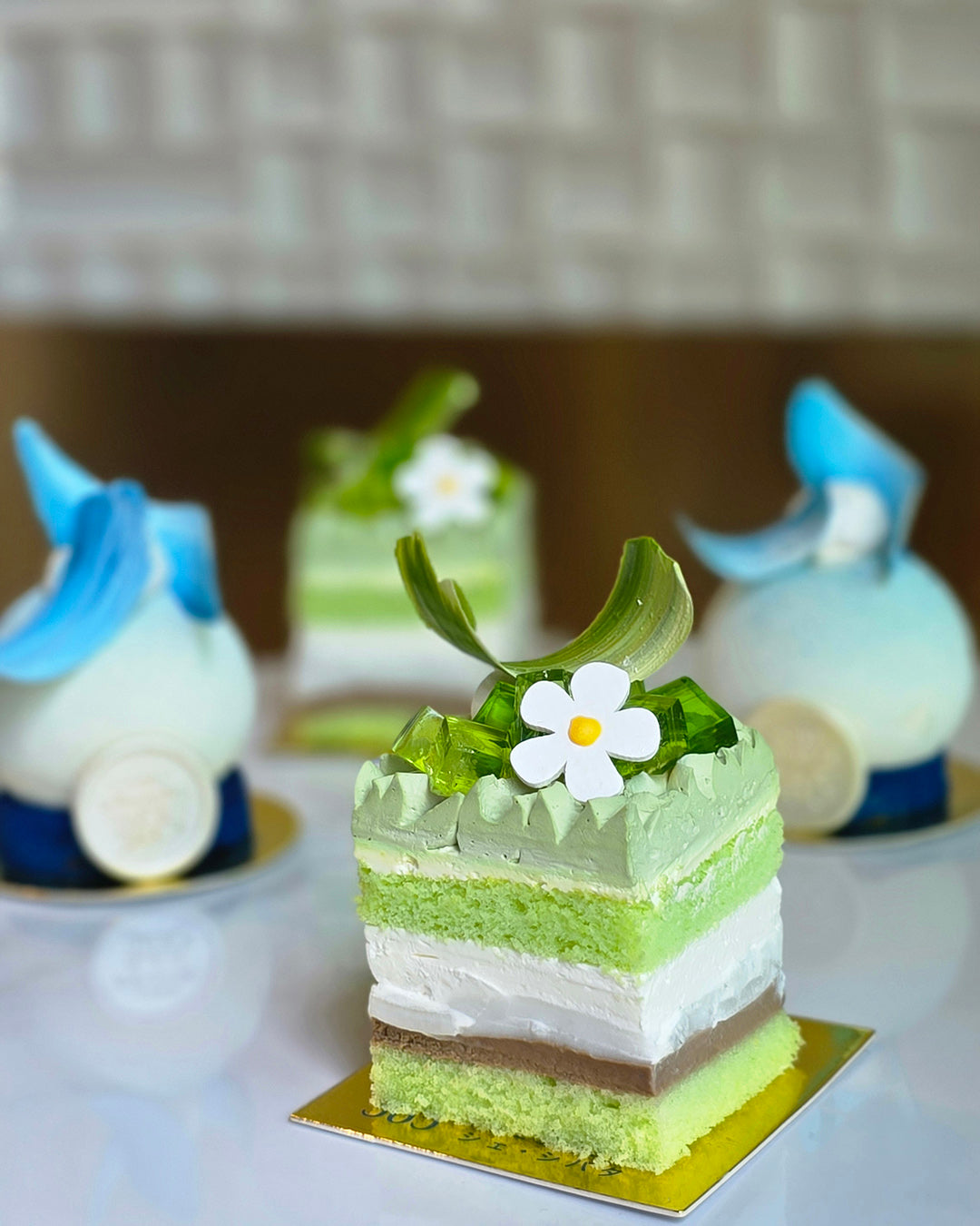 A square layered cake with green, white, and brown sponge and cream layers, topped with a white flower and green leaves, displayed on a gold square base. Two decorative blue and white dessert pieces are blurred in the background on a white surface. from Chez shibata365
