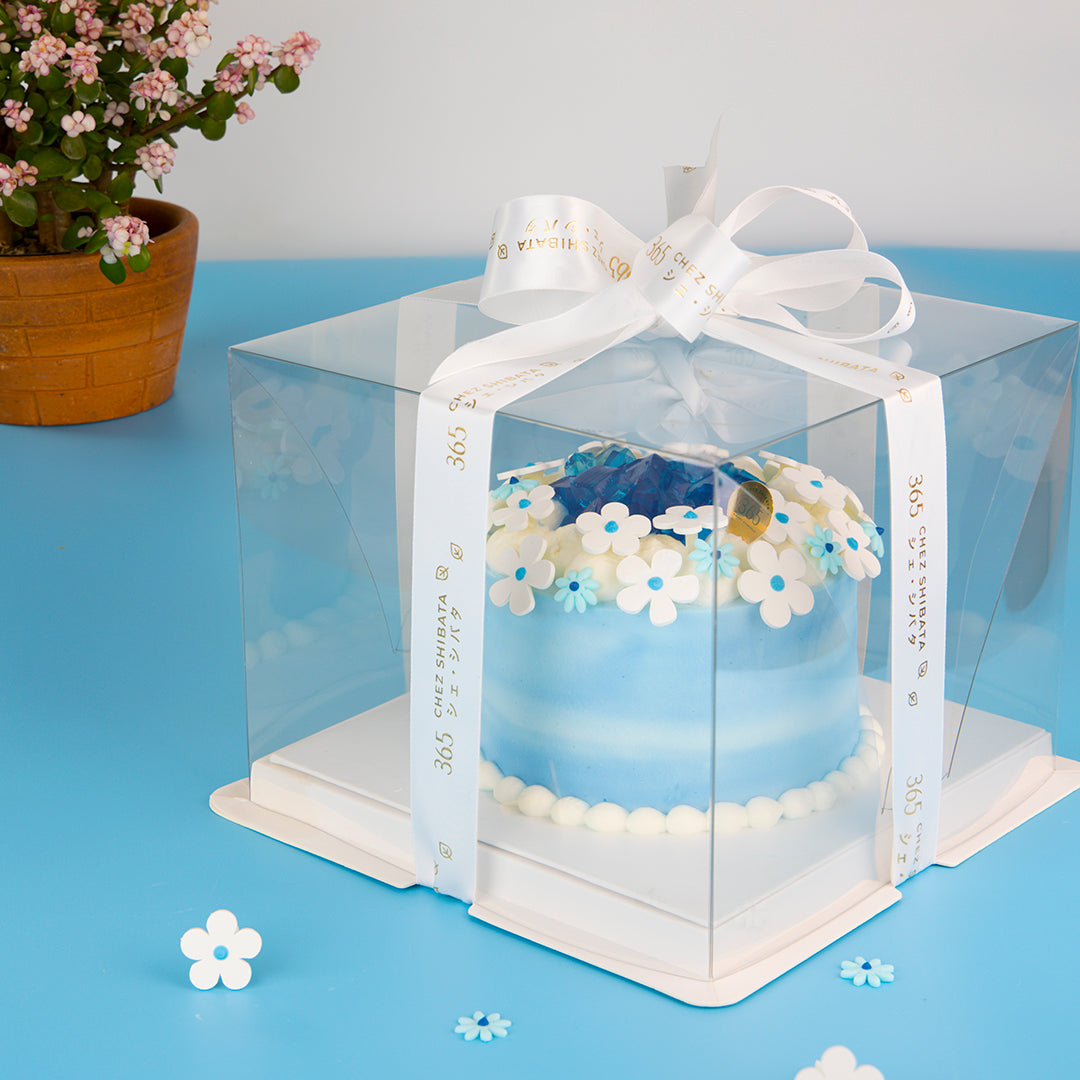 Clear gift box with a white ribbon on a blue surface with flowers. Coconut cake from Chez Shibata365