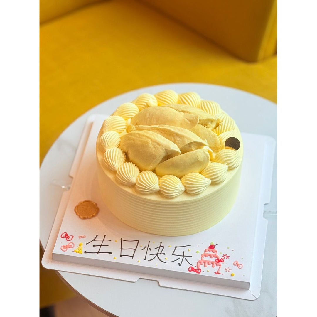 A round yellow birthday cake with piped cream decorations on top, presented on a white cake board with Chinese text that reads &