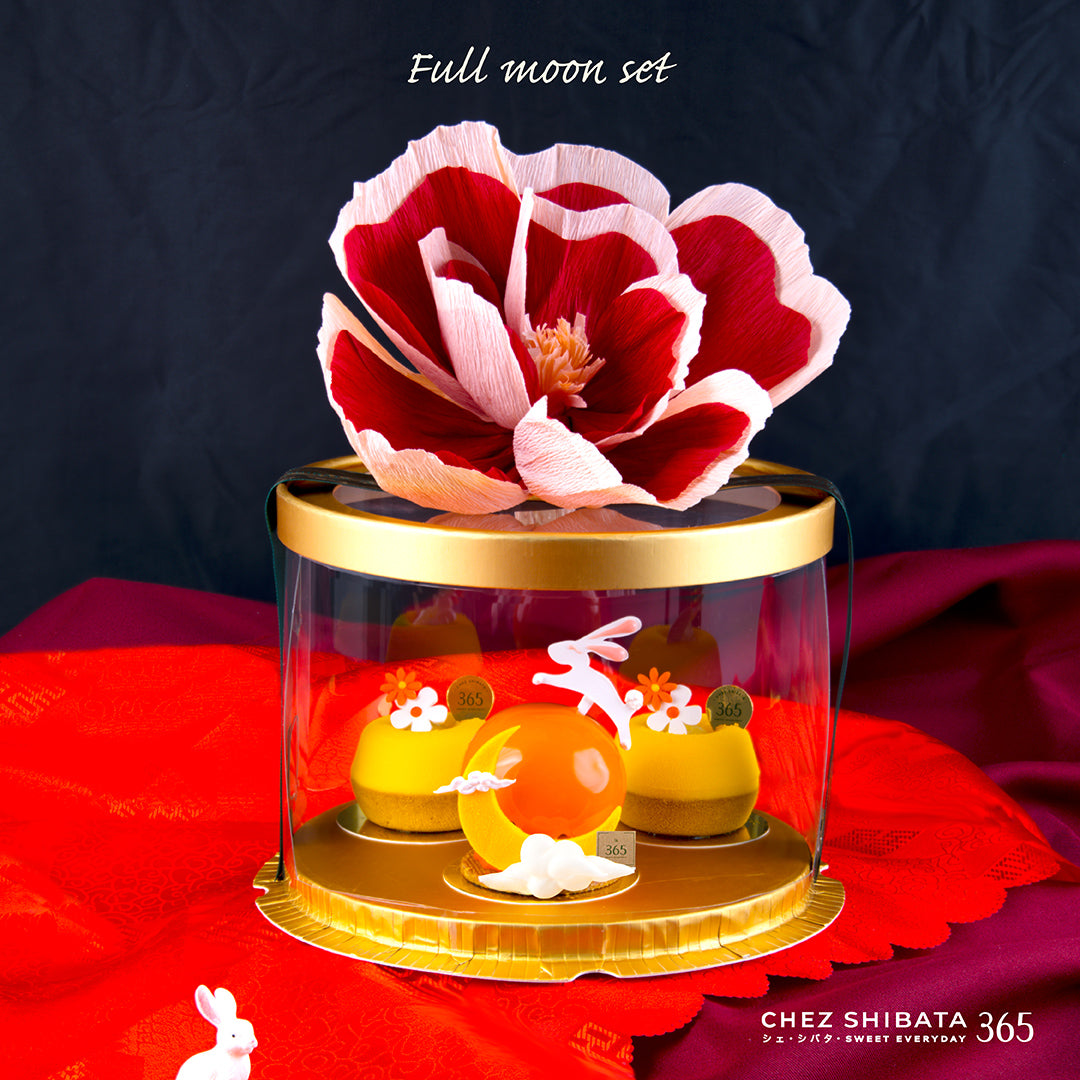 Full Moon Set by Chez Shibata 365 — a glossy orange mousse cake and mini yellow cakes in a clear round box, topped with a red-and-white paper flower.
