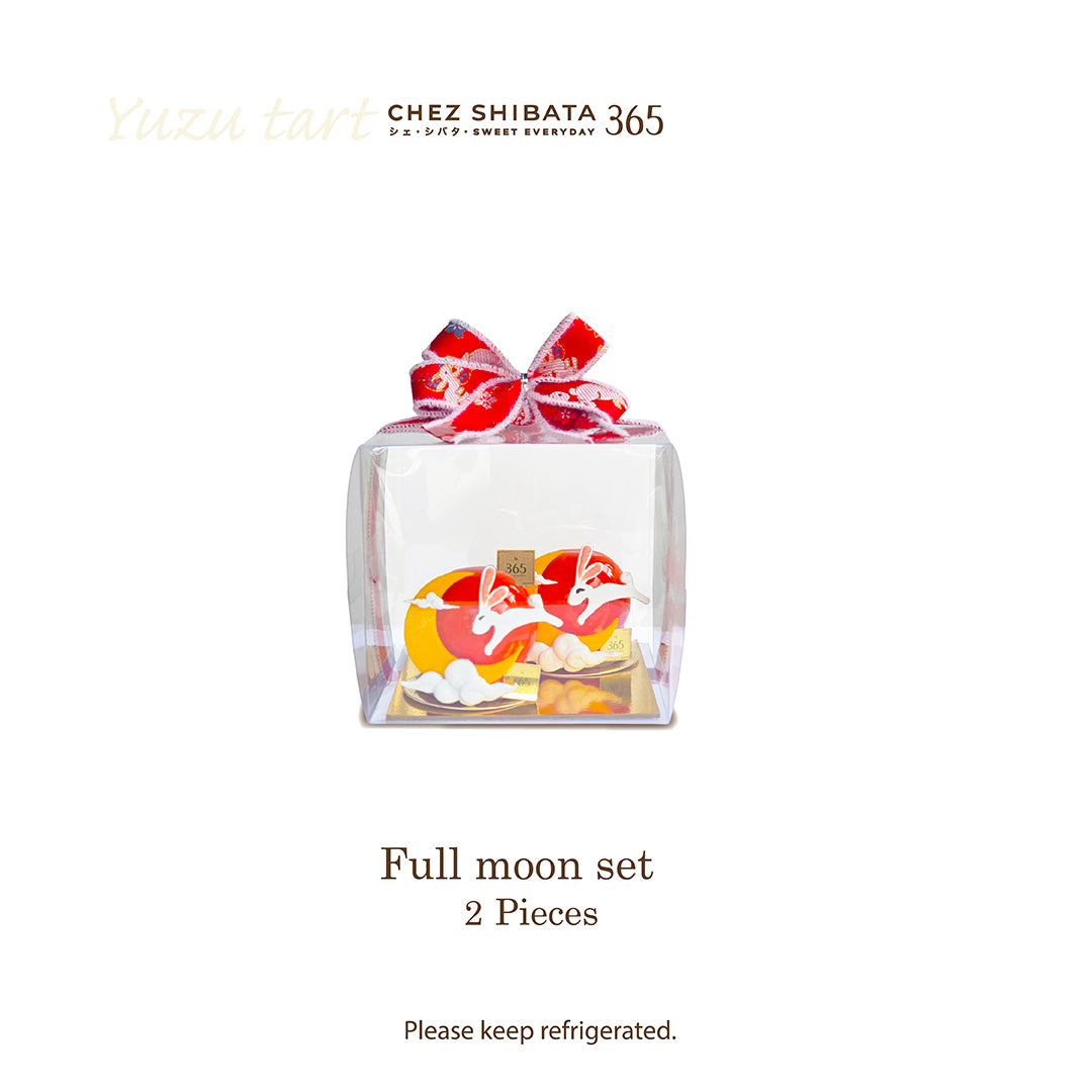 A clear square gift box containing a full moon-shaped pastry set with red and cream icing, tied with a red patterned bow. The image includes the brand text &