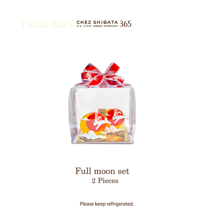 A clear square gift box containing a full moon-shaped pastry set with red and cream icing, tied with a red patterned bow. The image includes the brand text &