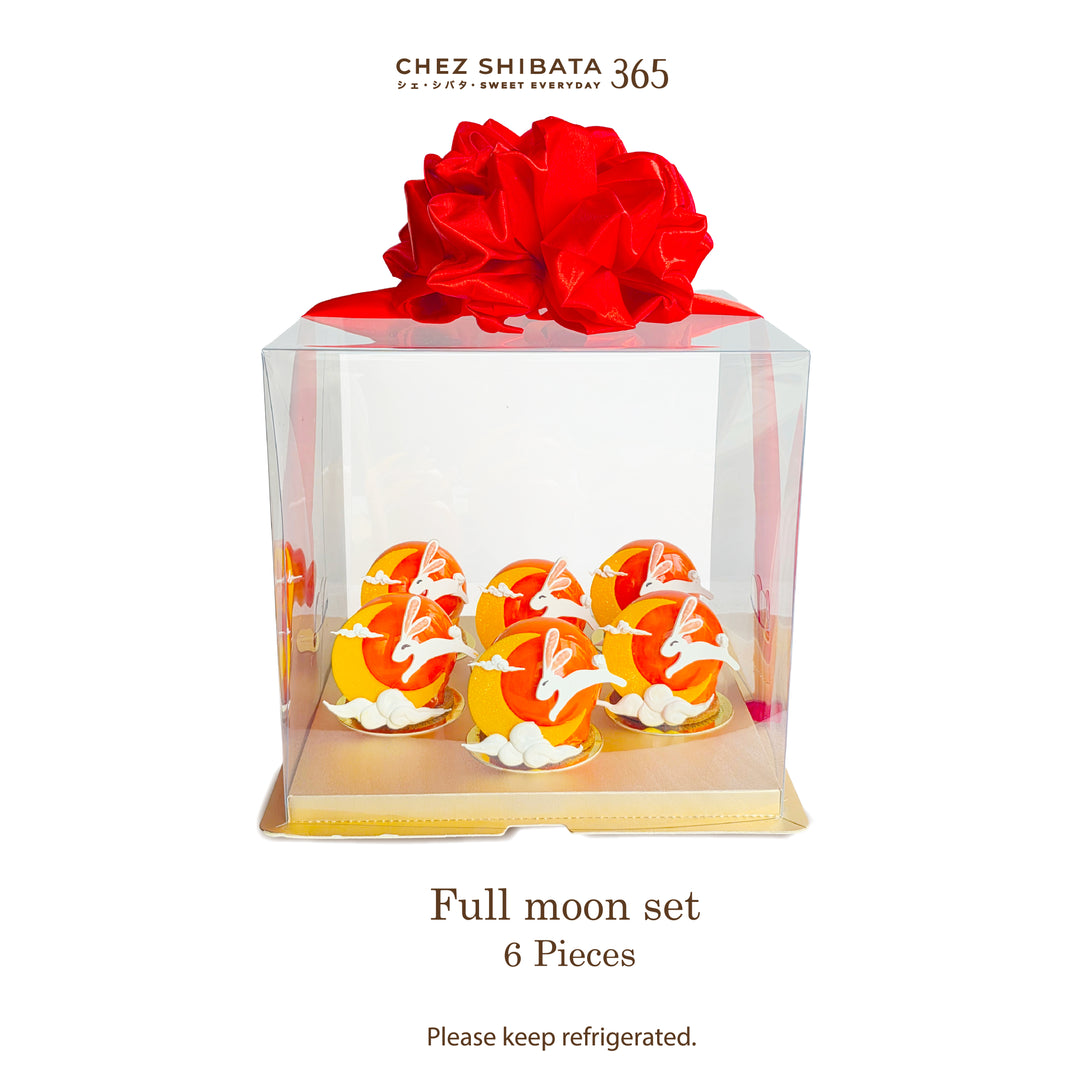 A clear square gift box containing six decorated moon cake pastries with orange and yellow fillings and white rabbit motifs, topped with a large red bow. The brand name Chez Shibata 365 appears at the top, and text at the bottom reads &