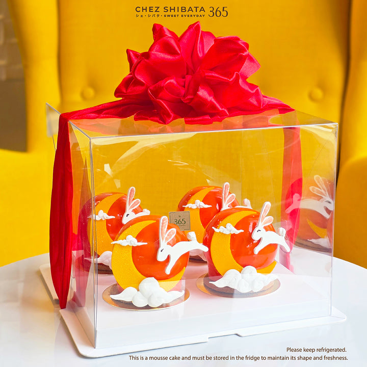 A clear square cake box with a large red satin bow and red ribbon, containing four decorated mousse cakes with orange moon shapes, white rabbit silhouettes, and white clouds. The box sits on a white surface against a bright yellow background. Moon cake