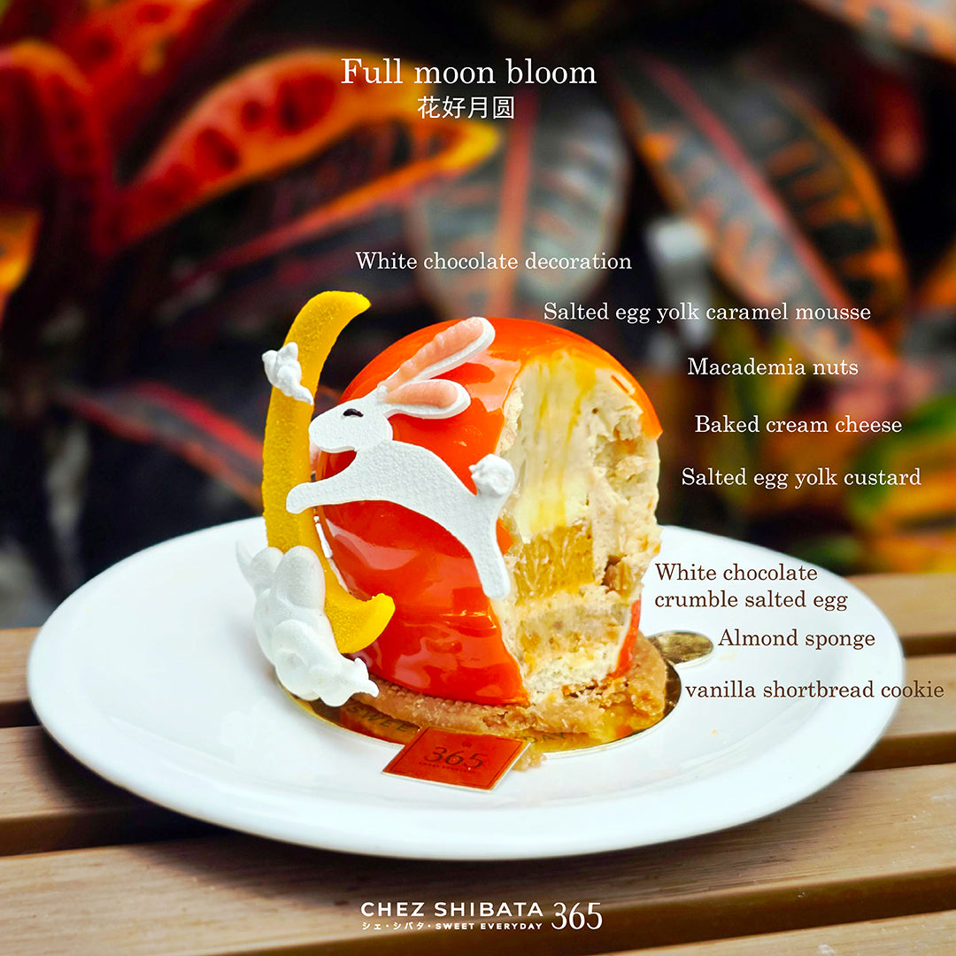 A beautifully decorated dessert from Chez Shibata365 on a white plate, featuring a glossy orange-red glaze, white rabbit decoration, yellow crescent moon, and cream-colored filling. The background is a blurred warm-toned setting with text overlays detailing the ingredients of the dessert.