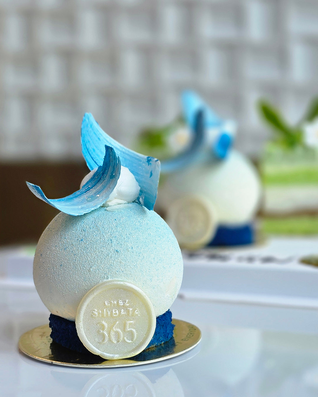 A close-up of a round white and pale blue dessert or pastry on a small gold plate, decorated with a curled light blue icing piece and a round white emblem reading 'chez shibata 365'. A second similar dessert is blurred in the background on a white surface. Cake near me