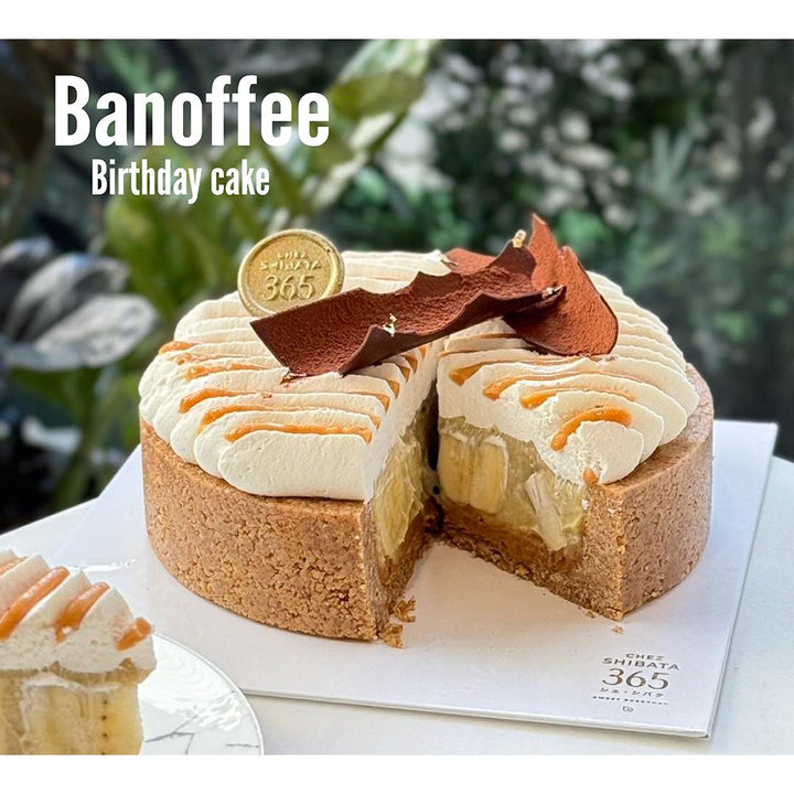 A Banoffee birthday cake from Chez Shibata 365, featuring a thick biscuit crust, layers of fresh banana and caramel, topped with piped whipped cream and chocolate decorations, displayed on a white base with a slice served on a plate nearby.