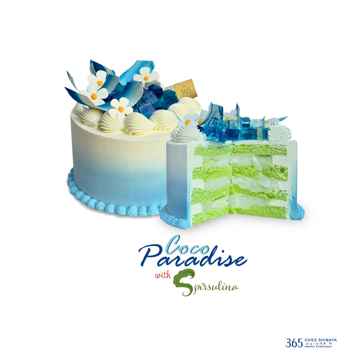 A blue and white tiered cake decorated with seashells, blue flowers, and white chocolate details, shown with a slice cut out to reveal green and white layers. The text &