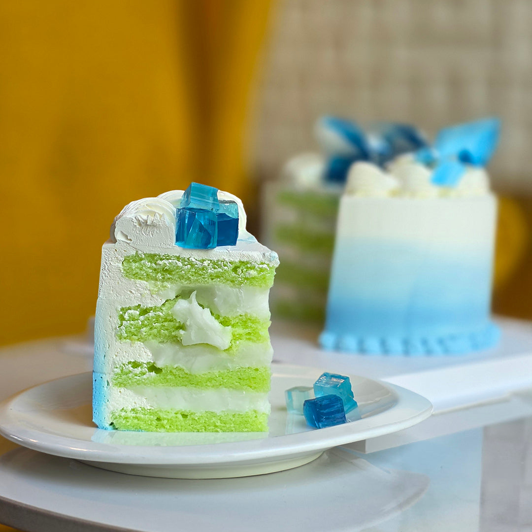 A slice of layered cake on a white plate, featuring light green sponge layers, white cream, and blue gelatin cubes, with a whole matching cake blurred in the background. Bangkok cake by Chez shibata365