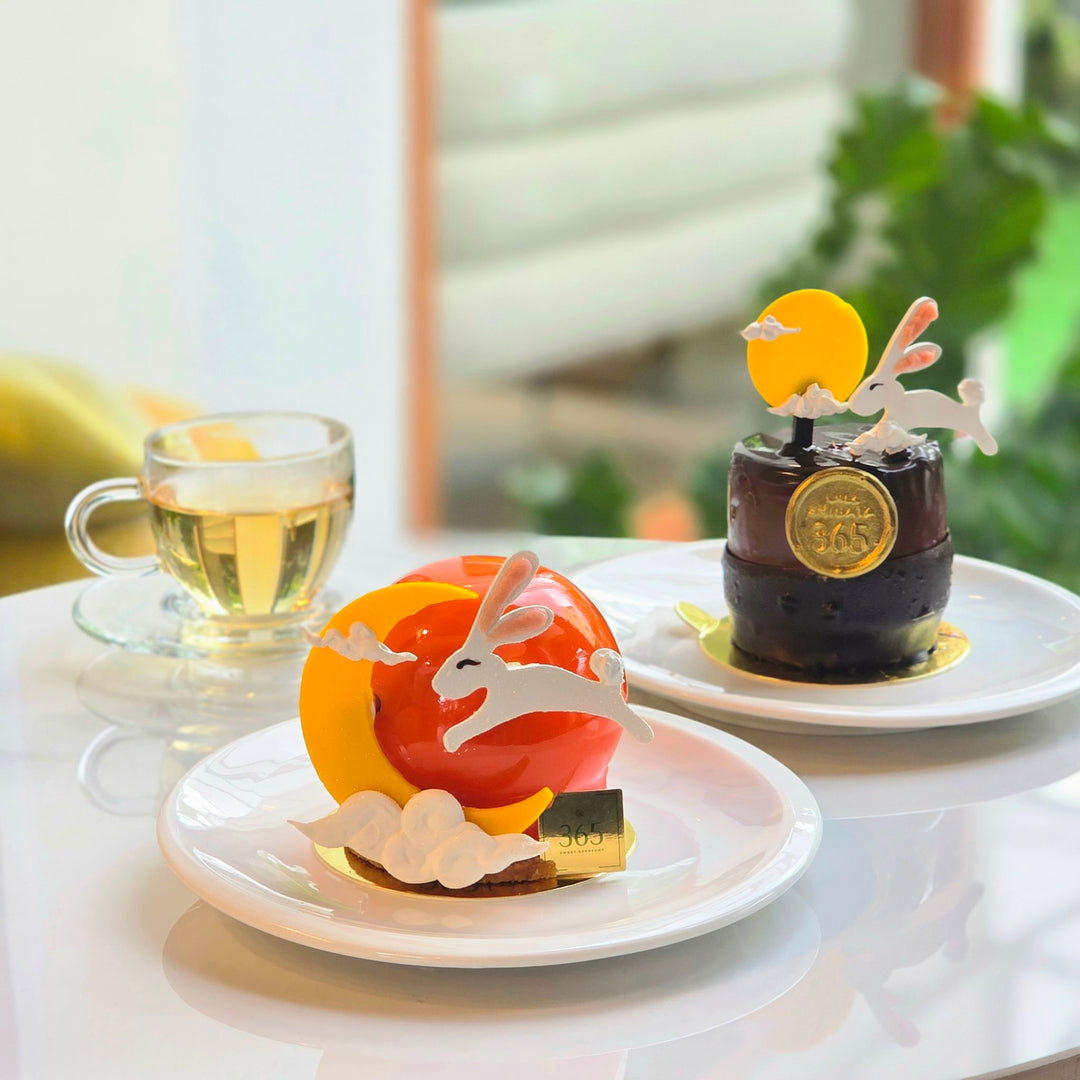 Two decorative desserts on white plates and a glass cup of tea in a bright indoor setting. One dessert is glossy red-orange with white rabbit-shaped decorations, and the other is a dark chocolate cake with a gold medallion and white rabbit decoration. cake from Chez Shibata365